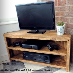 May include: A corner TV stand made of natural oak wood with two shelves. The stand has a rustic look and is perfect for displaying a TV, gaming console, and other electronics. The stand is shown with a TV, a gaming console, and a receiver on it. The stand is also shown with two remote controls on the top shelf. The stand is sitting on a tiled floor. "NATURAL OAK" is written on the floor in front of the stand. "Read the least the last 5-10 feedbacks please!" is written on the floor in front of the stand.