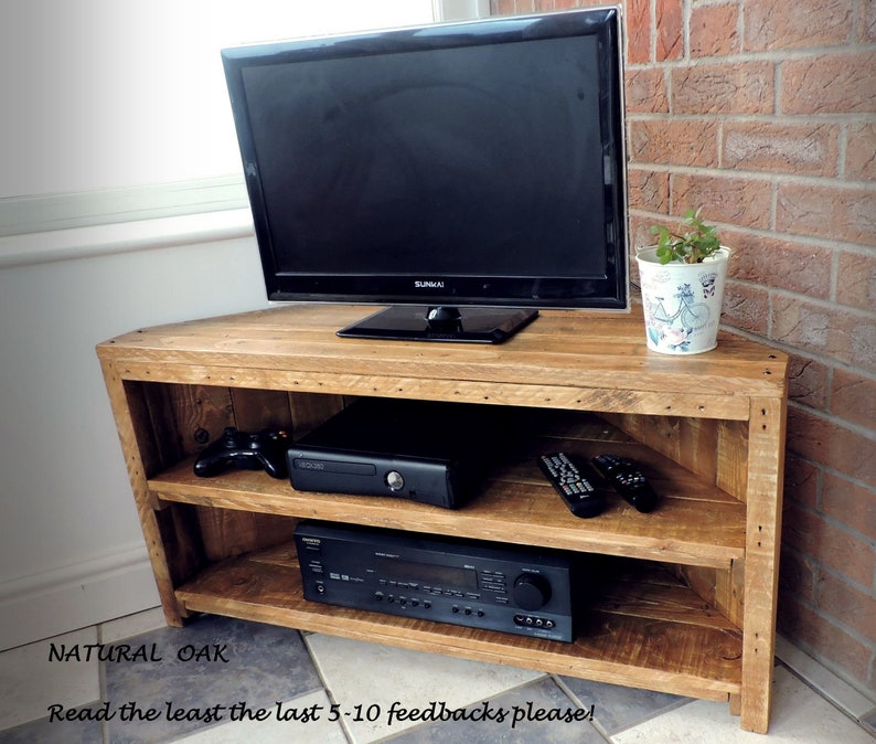 May include: A corner TV stand made of natural oak wood with two shelves. The stand has a rustic look and is perfect for displaying a TV, gaming console, and other electronics. The stand is shown with a TV, a gaming console, and a receiver on it. The stand is also shown with two remote controls on the top shelf. The stand is sitting on a tiled floor. "NATURAL OAK" is written on the floor in front of the stand. "Read the least the last 5-10 feedbacks please!" is written on the floor in front of the stand.