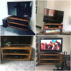 May include: A corner TV stand made of reclaimed wood with two shelves. The stand is shown with a TV on top and a sound bar on the bottom shelf. The stand is a dark brown color and has a rustic look.