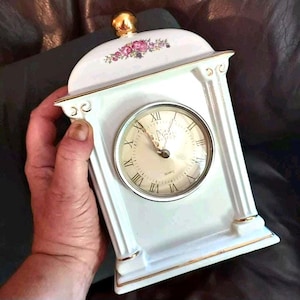 May include: A white ceramic mantel clock with gold trim and a floral design. The clock face has Roman numerals and the word "Quartz". The clock is approximately 6 inches tall.