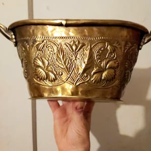May include: A vintage brass planter with ornate floral and leaf detailing. The oval-shaped container has two handles and a hammered texture, showcasing a warm, golden hue. The embossed design adds a touch of elegance to this decorative piece.