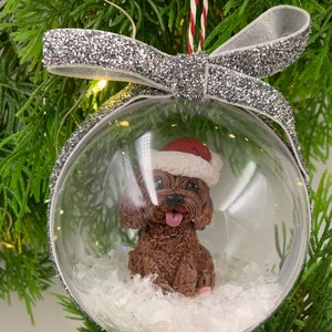 May include: A clear glass Christmas ornament with a brown dog wearing a red Santa hat inside. The ornament is filled with white faux snow and is decorated with a silver glitter ribbon.