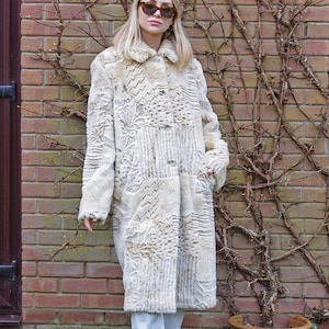 May include: A long, cream-colored faux fur coat featuring a patchwork design with various textured patterns, including zigzags and floral motifs. The coat has a classic collar, long sleeves, and a button-front closure, worn over light-colored trousers.