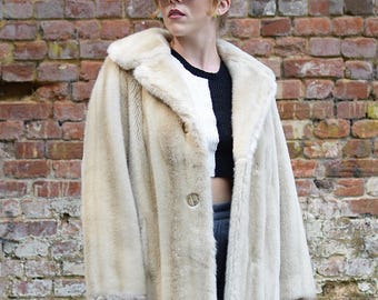 May include: A cream-colored faux fur coat with a wide collar and a single button closure. The coat is worn over a black and white color-blocked ribbed top and dark gray trousers. The ensemble is accessorized with dark-tinted sunglasses.
