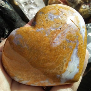 May include: A heart-shaped polished agate stone with a brown, orange, and white color pattern. The stone has a smooth, polished surface and a natural, organic shape.