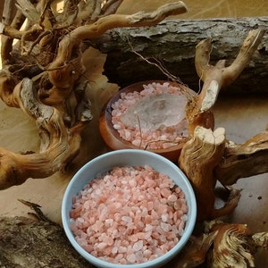 May include: Two bowls of pink Himalayan salt crystals, one with a clear quartz crystal, on a wooden surface with a natural branch background.