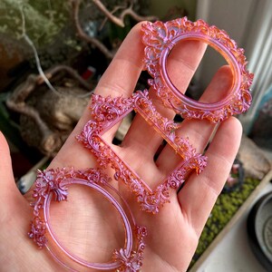 May include: Three ornate, decorative frames in shades of pink and orange are held in a hand. The frames are in oval, round, and diamond shapes, each with intricate floral and scrollwork detailing. The frames appear to be made of a translucent material.