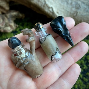 May include: Three miniature, handcrafted bottles and a raven skull. One bottle is shaped like a hand, another has a gold-speckled stopper, and the third is a small black raven skull. The bottles have a unique, aged appearance.