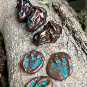 May include: Three small three-dimensional dinosaur skull figurines and three flat oval-shaped tokens featuring embossed dinosaur footprint designs. Each piece has a metallic copper finish with bright turquoise accents in the recessed areas.