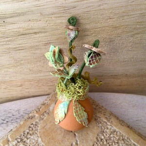 May include: A small, terracotta-colored pot with a green moss-like material around the top. The pot is decorated with a green and brown patterned design.  The pot contains a small arrangement of green and brown paper flowers.