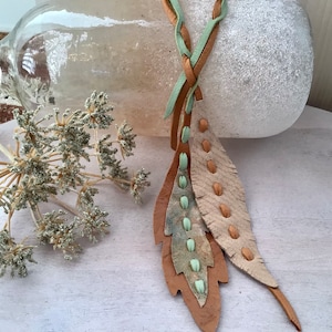 May include: A long leather necklace with two feather-shaped pendants. The necklace is braided with brown and green leather and has small brown beads along the edges of the feathers.