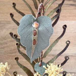 May include: A gray leather leaf pendant with a brown leather strap and a small, round, light brown bead. The pendant is hanging from a branch with small, round, brown buds.