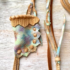 May include: A small, light green leather pouch with a brown leather trim and fringe. The pouch has a scalloped edge and is decorated with small, brown leather buttons.