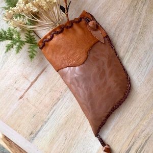May include: A brown leather pouch with a decorative stitched edge and a small leather flap. The pouch is hanging from a wooden surface and has a dried flower arrangement inside.