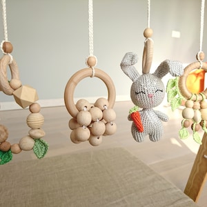 Op de afbeelding: Een houten babymobiel met vier hangende speelgoedjes. De speelgoedjes omvatten een ring van houten kralen met groene gehaakte bladeren, een ring van houten kralen met een houten ring, een gehaakt grijs konijn dat een wortel vasthoudt, en een ring van houten kralen met groene gehaakte bladeren.