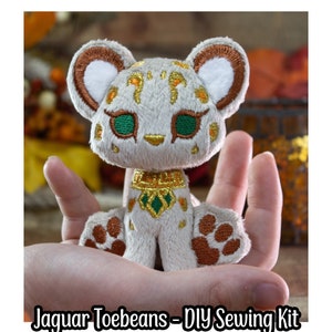 Puede incluir: Un jaguar de peluche blanco con manchas marrones y detalles dorados. El jaguar tiene ojos verdes y un collar dorado. El juguete de peluche está siendo sostenido en una mano. Jaguar Toebeans - Kit de costura DIY