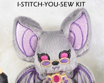 May include: A gray plush bat with purple wings and ears, wearing pink glasses and a gold and purple vest with a pink gem. The bat is sitting on a white surface with spools of thread and scissors.