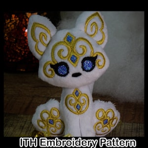 May include: White plush toy fox with gold embroidery and blue glitter eyes. The fox is sitting on a brown surface.