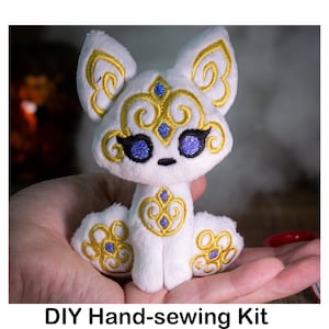 May include: DIY hand-sewing kit featuring a white plush fox with gold embroidery and blue glitter eyes. The fox is sitting on a hand.