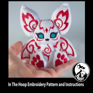 Könnte beinhalten: Weißes Plüschtier mit roter Stickerei in Form eines Fuchses mit großen Ohren. Das Spielzeug sitzt in einer Hand. Der Text "In The Hoop Embroidery Pattern and Instructions" befindet sich am unteren Rand des Bildes.