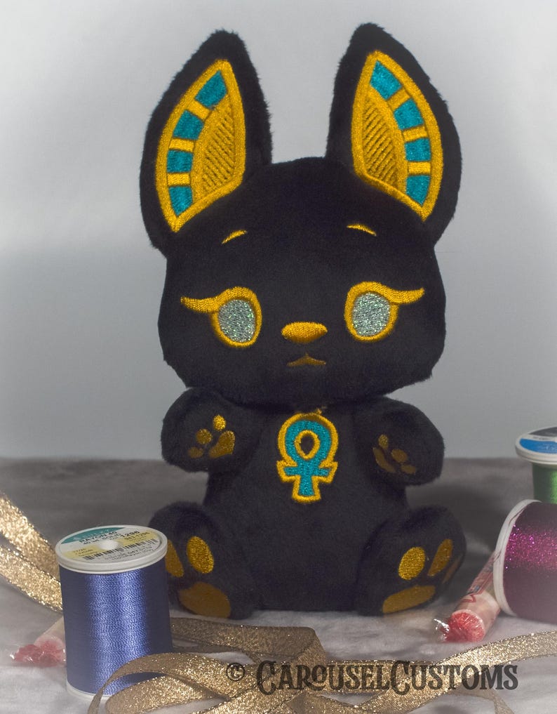 May include: Black plush toy shaped like a cat with gold and teal embroidered details. The toy has a large Ankh symbol on its chest. The toy is sitting on a white surface with gold ribbon and spools of thread.
