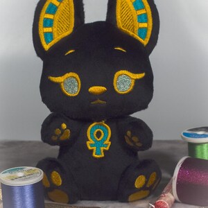 May include: Black plush toy shaped like a cat with gold and teal embroidered details. The toy has a gold ankh symbol on its chest. The toy is sitting on a table with gold ribbon and spools of thread.