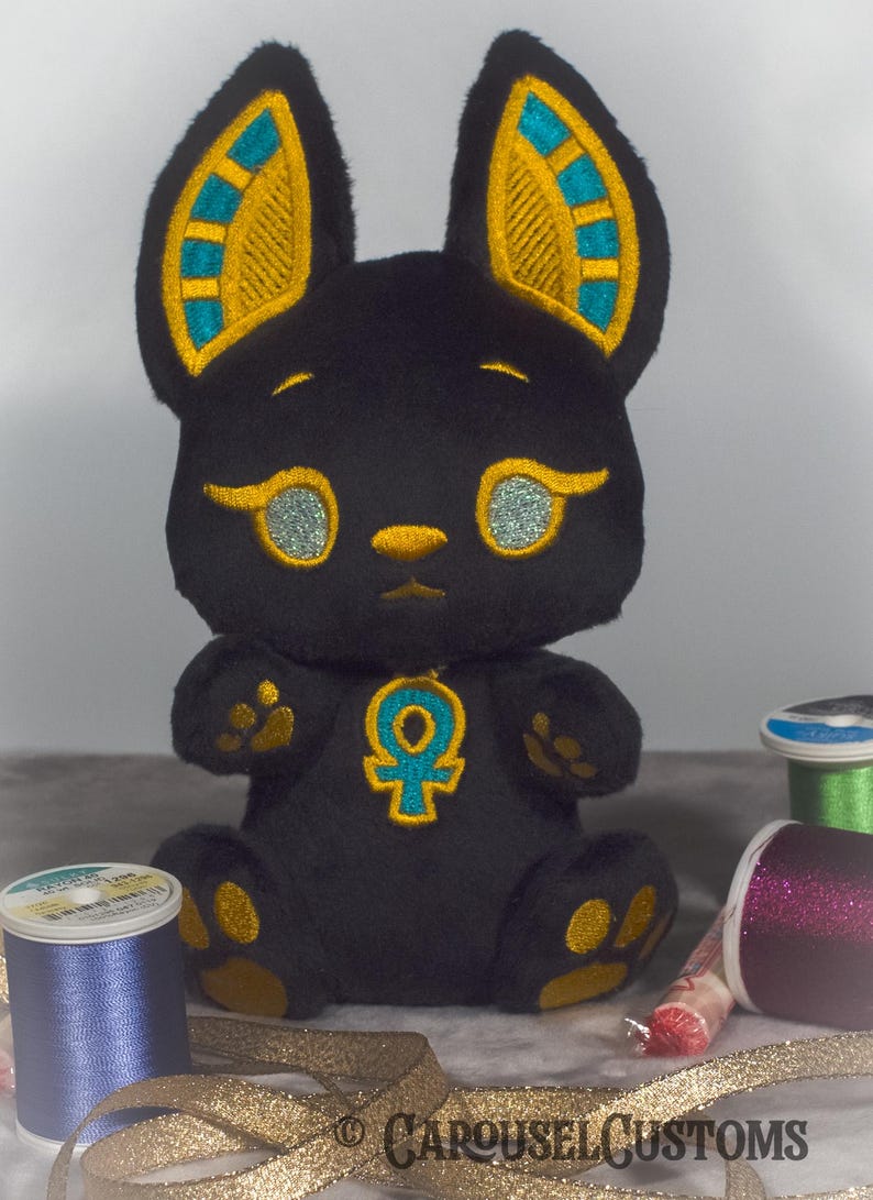 May include: Black plush toy shaped like a cat with gold and teal embroidered details. The toy has a gold ankh symbol on its chest. The toy is sitting on a table with gold ribbon and spools of thread.
