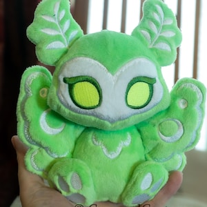 May include: A green plush moth toy with white and silver embroidered details. The toy features large eyes and leaf-shaped antennae. The wings and body are a vibrant green colour. The toy is soft and cuddly.