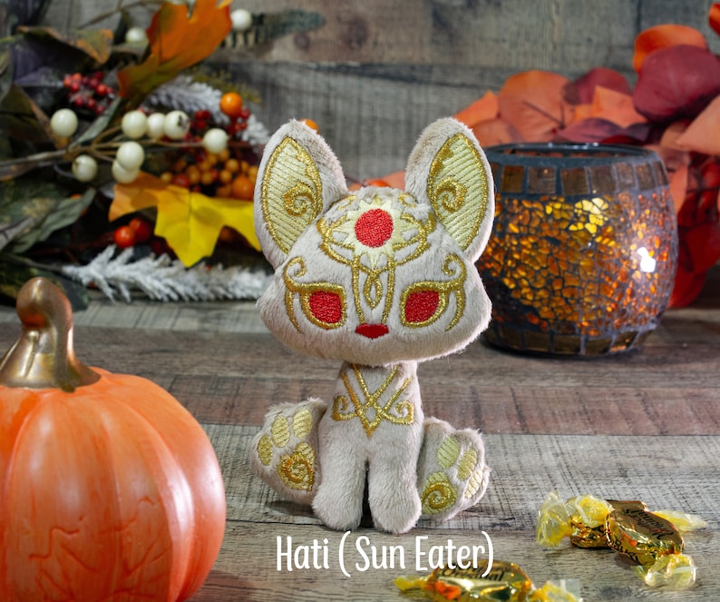 May include: A plush toy of a white wolf with gold embroidery and a red circle on its forehead. The wolf is sitting on a wooden surface with a pumpkin and other fall decorations in the background. The text "Hati (Sun Eater)" is written below the wolf.