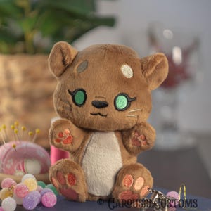 May include: A brown plush toy with a cat-like face, green eyes, and pink paw prints. The toy has a white belly and is sitting on a blue surface. The toy has a key chain attached to it.