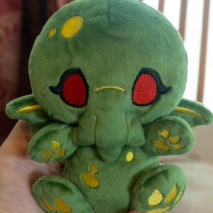 May include: A plush green creature with red eyes and yellow accents. The stuffed animal has small wings and is holding a pose. The plush toy is a unique and whimsical design.