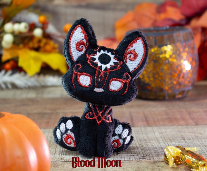 May include: Black plush toy with red and white embroidery depicting a stylized animal with a sun and moon design. The toy is sitting on a wooden surface with the text "Blood Moon" below it.