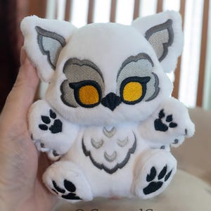 Könnte beinhalten: Ein weißes Plüsch-Eulen-Spielzeug mit gestickten grauen und gelben Details. Die Eule hat große gelbe Augen, graue Akzente und schwarze Pfotenabdrücke. Der Text "CarouselCustoms" befindet sich unten.