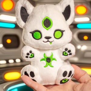 May include: A white plush toy cat with black ears and accents, and green eyes and markings. The cat has a green symbol on its chest and paw pads. The toy is held in a person's hand.