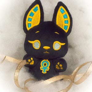 May include: Black plush toy shaped like a cat with gold and blue ears and a gold ankh symbol on its chest. The toy is sitting on a white fluffy surface with a gold ribbon.