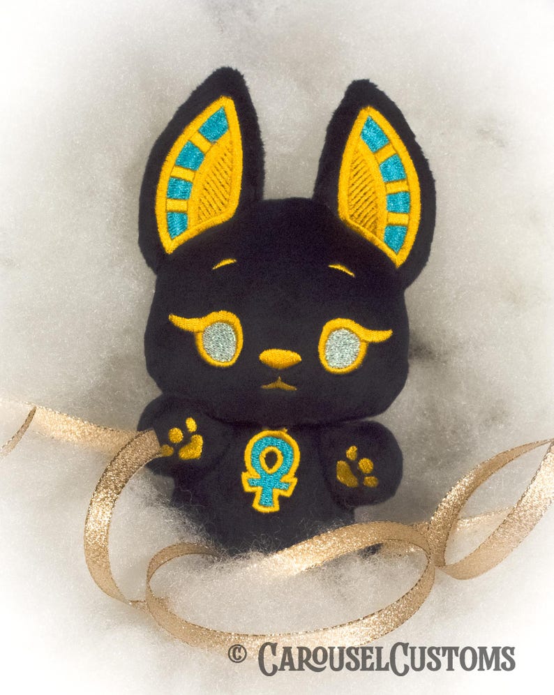 May include: Black plush toy shaped like a cat with gold and blue ears and a gold ankh symbol on its chest. The toy is sitting on a white fluffy surface with a gold ribbon.