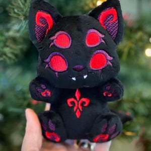 May include: A black plush toy shaped like a four-eyed cat with vibrant red embroidered eyes, inner ears, and paw pads. The creature features a red fleur-de-lis symbol on its chest and two small white fangs protruding from its mouth.