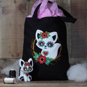 Puede incluir: Bolsa de tela negra con un zorro blanco bordado con un diseño de calavera de azúcar y detalles florales.