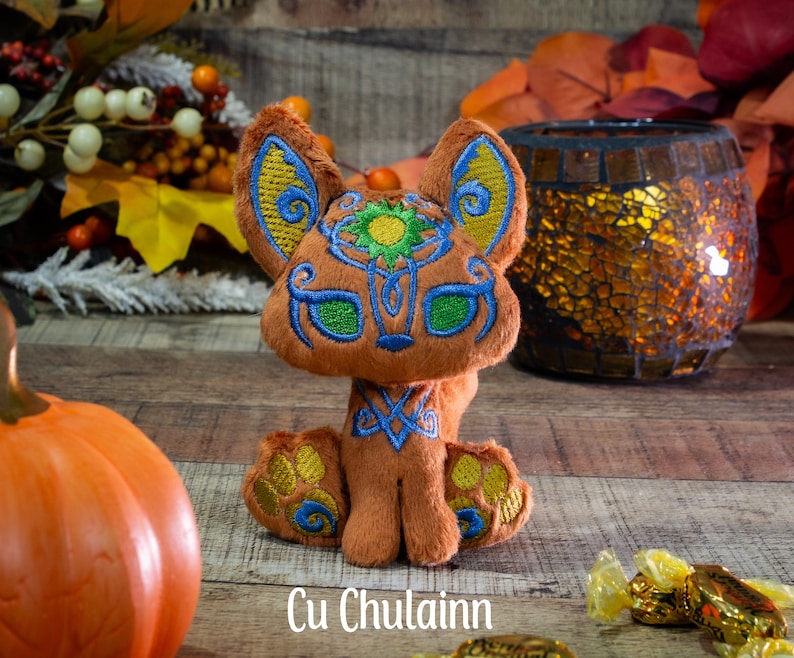 May include: A plush fox toy with orange fur, green eyes, and intricate embroidery details. The fox is sitting on a wooden surface with fall foliage and a pumpkin in the background. The text "Cu Chulainn" is visible below the fox.