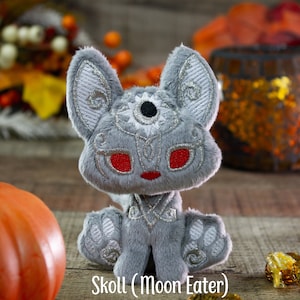 May include: A gray plush toy wolf with silver embroidery and red eyes. The toy is sitting on a wooden surface with a pumpkin and a decorative lantern in the background. The toy is labeled "Skoll (Moon Eater)".