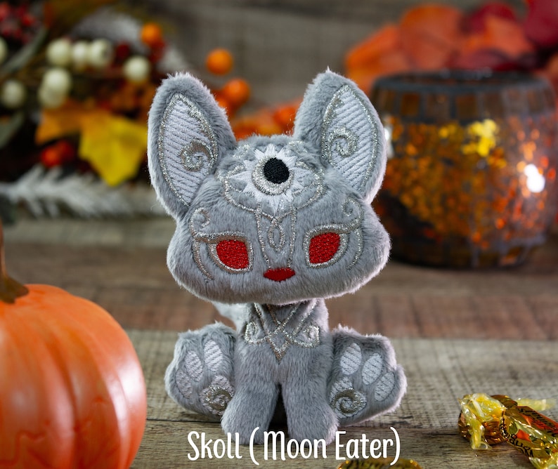 May include: A gray plush toy wolf with silver embroidery and red eyes. The toy is sitting on a wooden surface with a pumpkin and a decorative lantern in the background. The toy is labeled "Skoll (Moon Eater)".