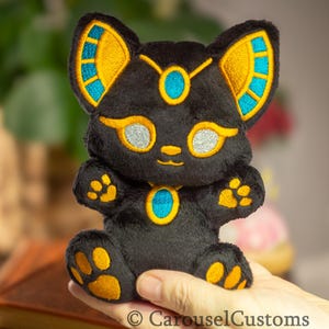 May include: Black plush cat toy with gold and blue embroidered details. The cat has a turquoise gem on its chest and gold paw prints on its feet.