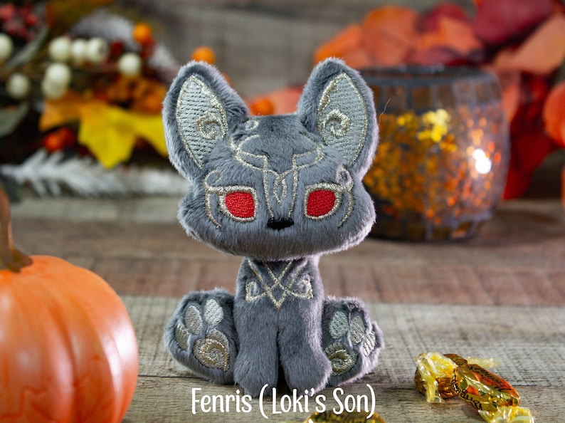 May include: A gray plush toy fox with red eyes and silver embroidery. The toy is sitting on a wooden surface with a pumpkin and candy in the background. The toy is labeled "Fennris (Loki's Son)".