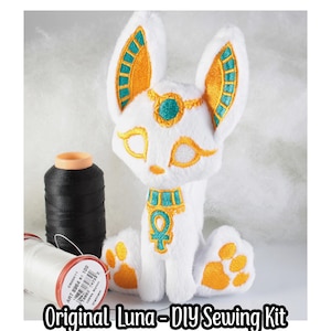 May include: A white plush toy cat with gold and teal embroidery. The cat is sitting with its paws outstretched. The cat has a gold collar with an ankh symbol. The toy is part of a DIY sewing kit called "Original Luna".
