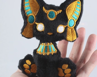 May include: Black plush cat with gold and turquoise embroidery. The cat is wearing a gold and turquoise collar and has gold paw prints. The cat is sitting on a hand. DIY Bast Embroidery Pattern.
