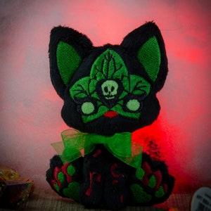 May include: Black plush cat with green embroidered leaves and a skull on its face. The cat is wearing a green ribbon bow and has red embroidered paw prints on its body.