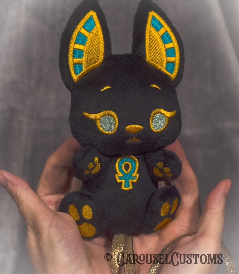 May include: Black plush toy shaped like an Egyptian deity Anubis with gold and turquoise embroidery. The toy has a golden Ankh symbol on its chest.