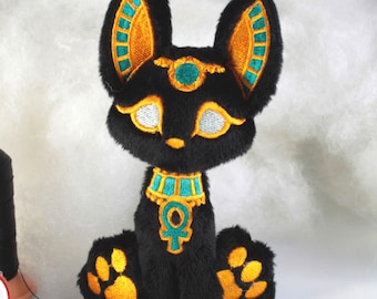 May include: Black plush toy of the Egyptian god Anubis, a jackal-headed deity, with gold and teal embroidery details. The toy is sitting with its paws visible.