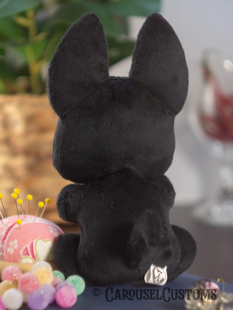 May include: Black plush toy in the shape of a dog sitting on a table. The toy has a small tail and a tag on its back that says "Carousel Customs".