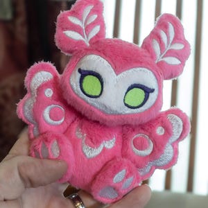 Puede incluir: Un peluche rosa con forma de criatura con orejas grandes, detalles blancos y ojos verdes. El peluche tiene detalles bordados en plata en las alas y los pies. El texto "CarouselCustoms" es visible en la parte inferior.